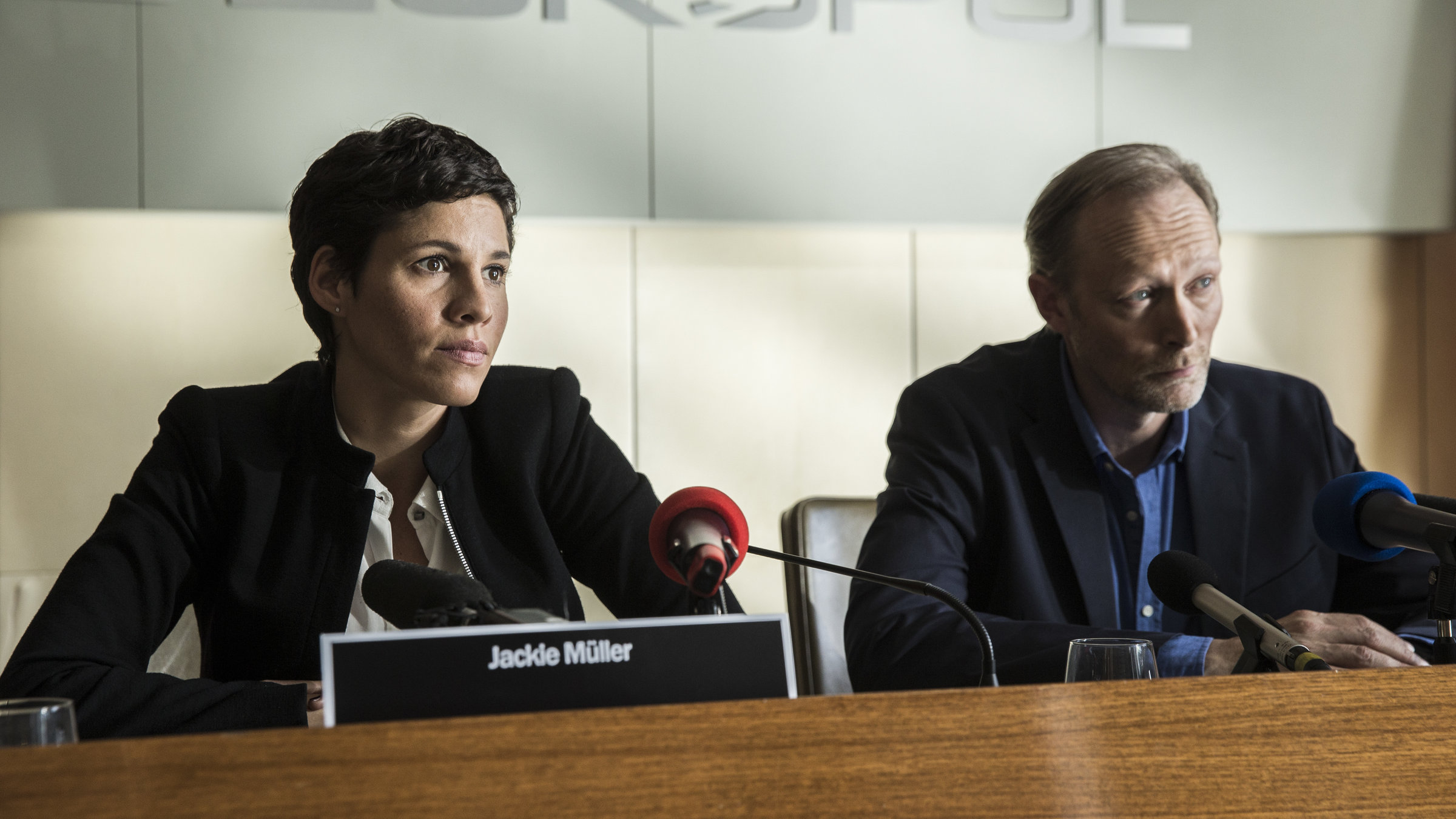 "Das Team (2)": Jackie Mueller (Jasmin Gerat) und Harald Bjørn (Lars Mikkelsen) sitzen nebeneinander an einem Tisch vor Mikrophonen.