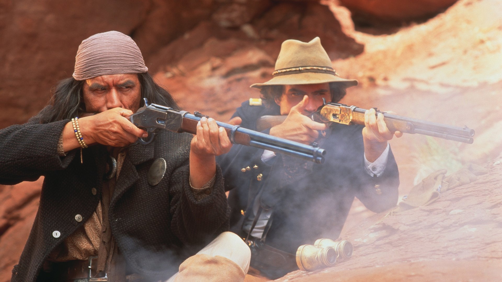 "Geronimo – Eine Legende": Apachen-Häuptling Geronimo (Wes Studi, links) in einem grauen Mantel, mit langen Haaren und Kopftuch, und Lt. Charles Gatewood (Jason Patric) in Soldatenuniform haben sich vor roten Felsen verschanzt und feuern aus ihren Gewehren.