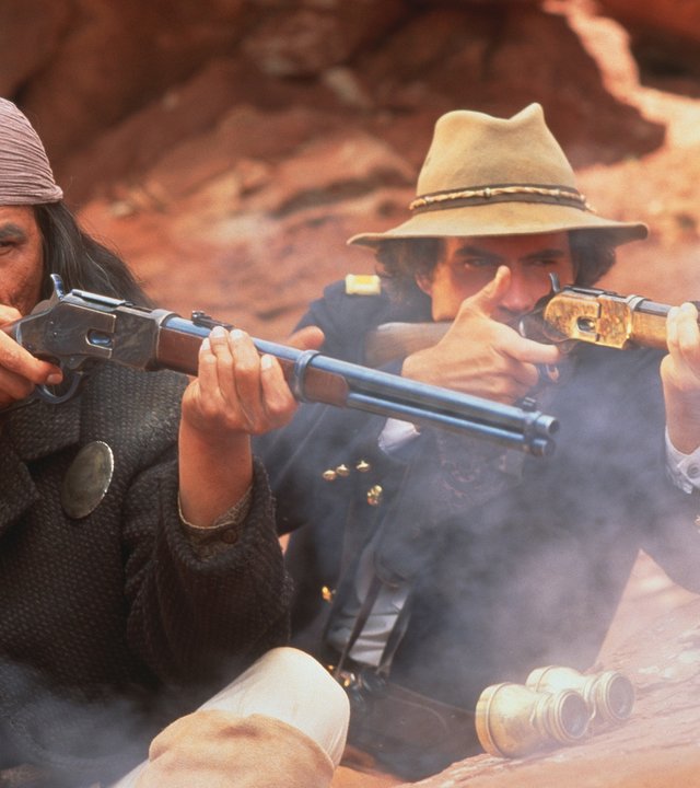 "Geronimo – Eine Legende": Apachen-Häuptling Geronimo (Wes Studi, links) in einem grauen Mantel, mit langen Haaren und Kopftuch, und Lt. Charles Gatewood (Jason Patric) in Soldatenuniform haben sich vor roten Felsen verschanzt und feuern aus ihren Gewehren.