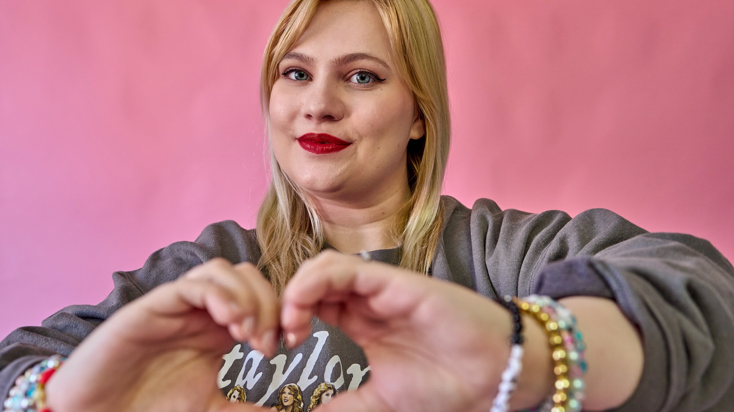 "37° - Being Swiftie. Kims Leben mit Taylor Swift": Kim Niehaus (26) steht vor einem rosa Hintergrund und trägt einen Swift-Pullover und Armbänder. Sie formt mit ihren Händen ein Herz, durch das "Taylor" zu lesen ist.