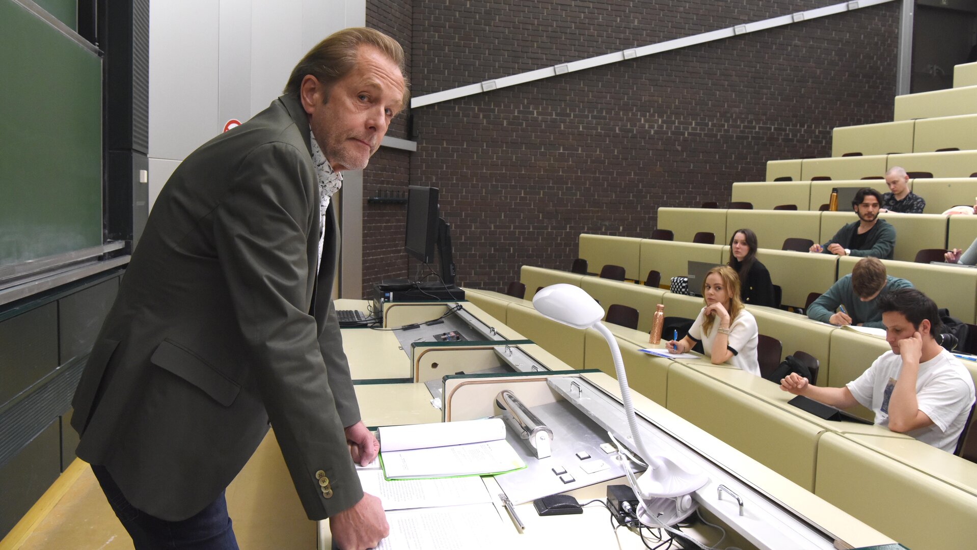 "SOKO Wien - Was wäre, wenn...?": Spurensicherer Franz Wohlfahrt (Helmut Bohatsch) steht an einem Pult im Hörsaal. Vor ihm sitzen Studenten (Komparsen), die sich Notizen machen.