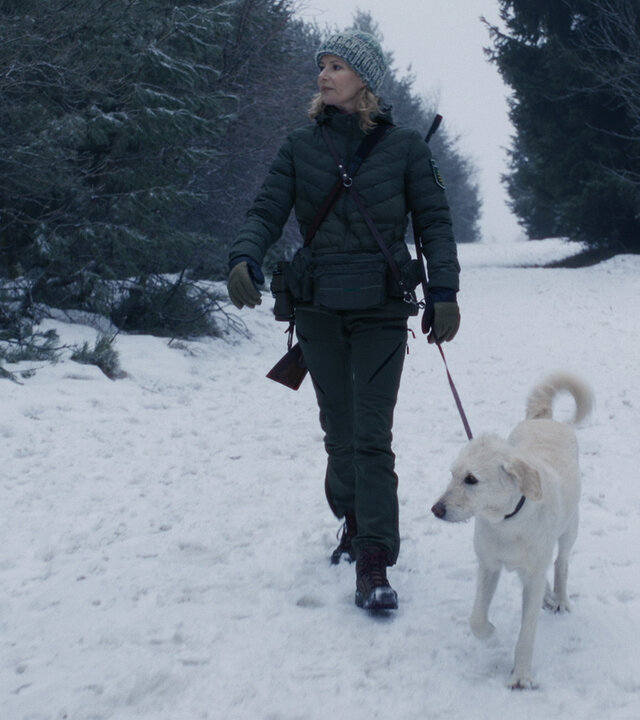 "Erzgebirgskrimi - Wintermord": Die Försterin Saskia Bergelt (Teresa Weißbach) läuft mit Hund Wolke einen zugeschneiten Waldweg entlang. Sie trägt ihr Gewehr auf ihrem Rücken.