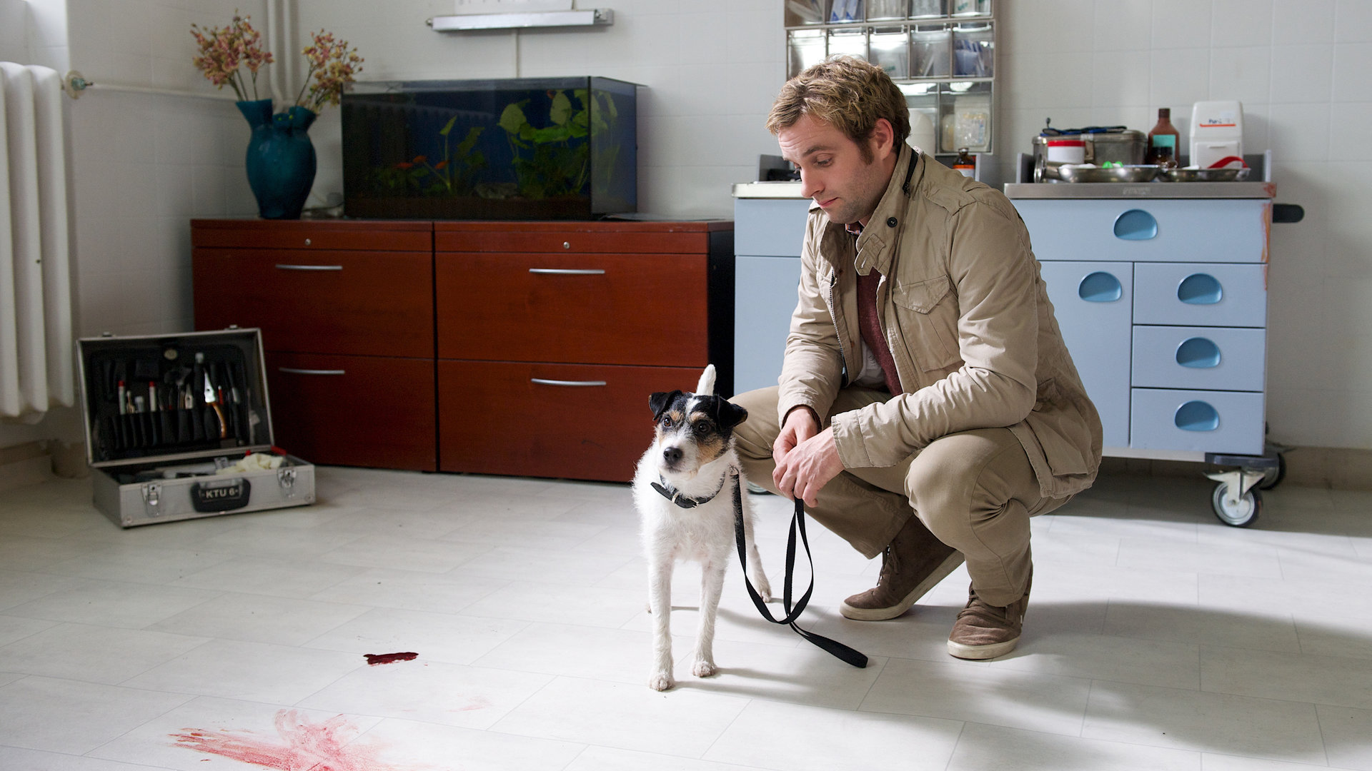"SOKO Wismar - Stummer Zeuge": Am Tatort, einem Wohnzimmer, hockt Nils Theede (Jonas Laux) mit einem Hund an der Leine. Im Hintergrund sind zwei Kommoden zu sehen sowie ein geöffneter Werkzeugkoffer und Blutflecken auf einem hellen Teppich.