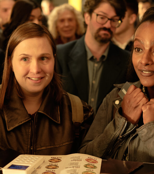 "Take the Money and Run - One Lie": Claire Walsh (Jana Julia Roth) und Abby (Yodit Tarikwa) stehen in einer Schlange an einem Tresen. Auf dem Tresen liegt ein OneCoin-Ordner, daneben steht ein Kartenlesegerät. Beide haben einen aufgeregten Gesichtsausdruck.