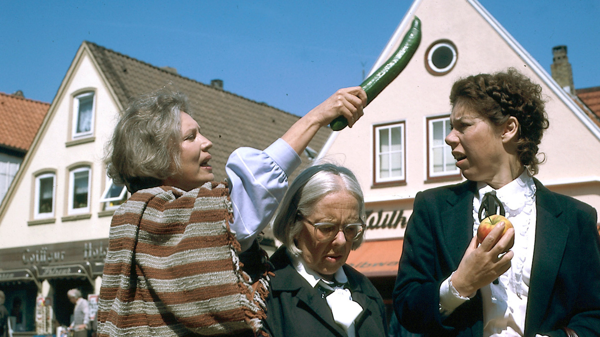 "Der Landarzt - Die Liebe, die Liebe": Olga Mattiesen (Antje Weisgerber) schlägt mitten auf einem Marktplatz Thea (Evelyn Hamann) mit einer Gurke auf den Kopf. Zwischen ihnen steht die irritierte Berta (Gerda Gmelin).