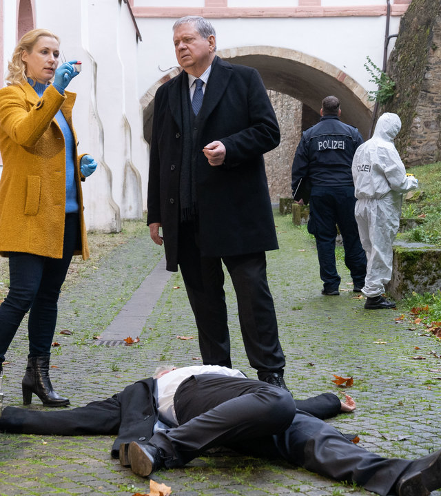 "Der Staatsanwalt - Erfolgreich, glücklich, tot": Dr. Judith Engel (Astrid Posner) und Oberstaatsanwalt Bernd Reuther (Rainer Hunold) stehen neben Noah Jansen (Richard Gonlag), der tot und mit verdrehten Beinen am Boden liegt. Dr. Engel, die blaue Handschuhe trägt, hält in ihrer rechten Hand etwas, auf dass sie und Reuther interessiert schauen. Um sie herum sind drei Polizisten und zwei Spurensicherer bei der Arbeit zu sehen.