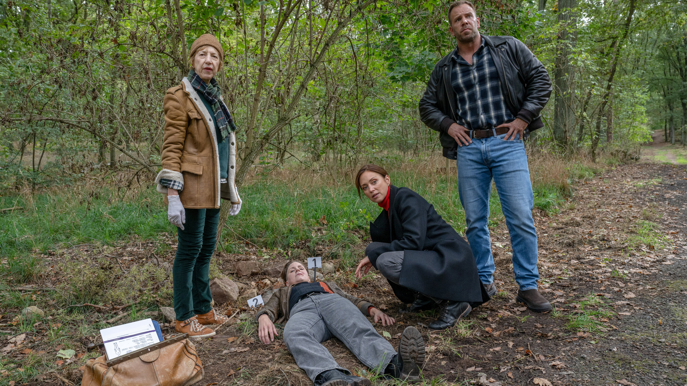 "SOKO Wismar - Karoline Undercover (1) - Die Giftschlange": Karoline Joost (Nike Fuhrmann), Dr. Helene Sturbeck (Katharina Blaschke) und Lars Pöhlmann (Dominic Boeer) stehen um die Leiche von Anja Schlüter (Kerstin Neutze), die auf einem Waldweg liegt.