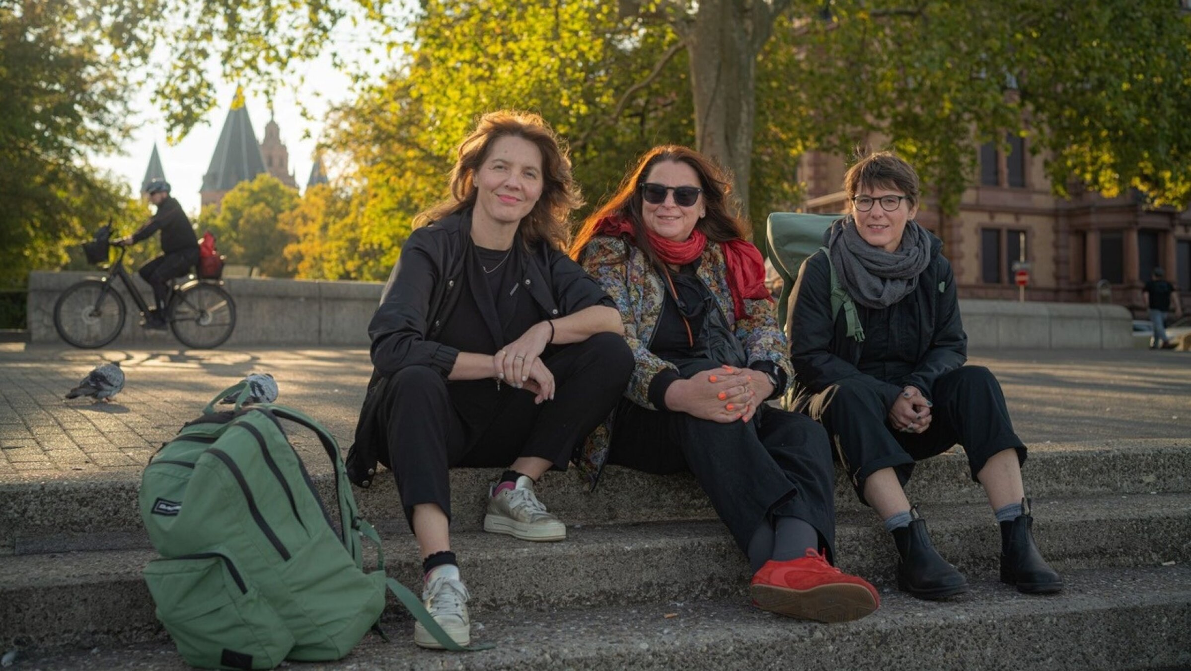 "Drei ostdeutsche Frauen bummeln durchs Land und prüfen die Schwerkraft der Verhältnisse": Peggy Mädler, Annett Gröschner und Wenke Seemann sitzen im Freien nebeneinander auf Steinstufen. Im Hintergrund sind die Türme des Mainzer Doms zu erkennen.