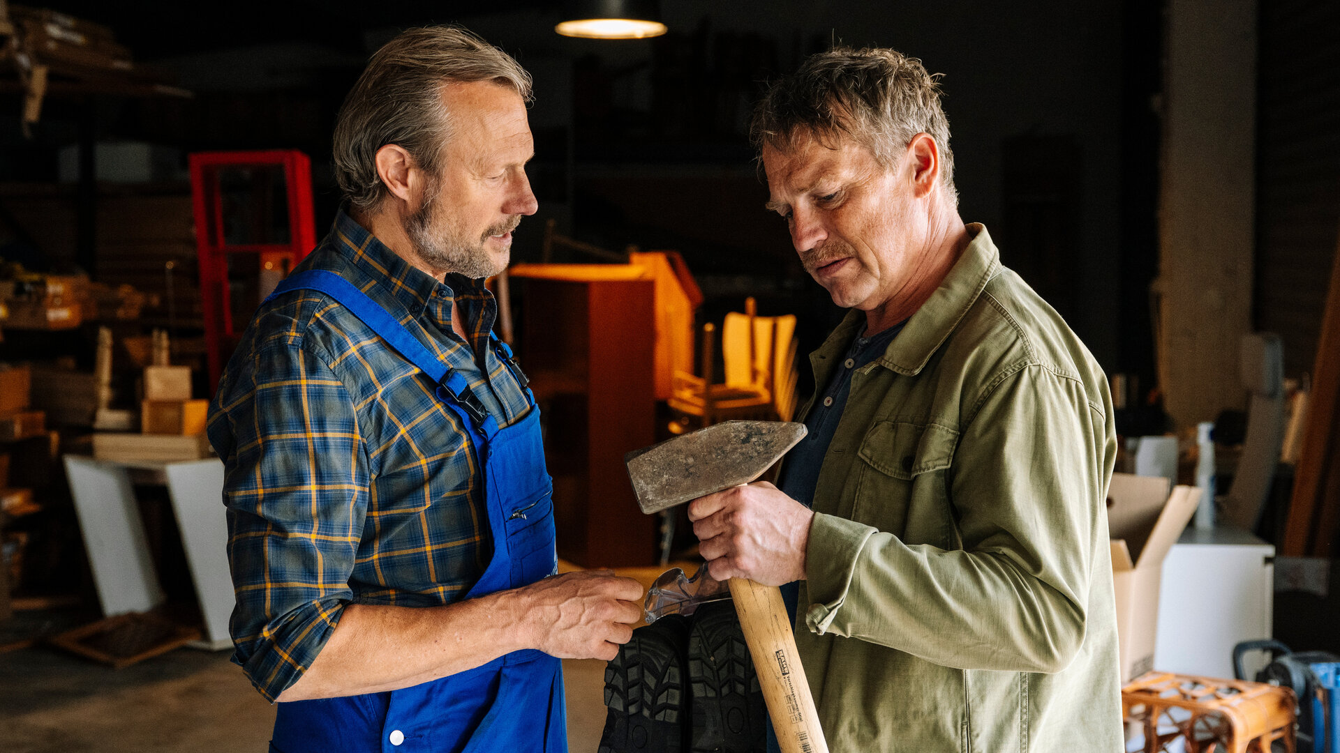 "SOKO Köln - Unvergessen": Matti Wagner (Pierre Besson) steht in einem Lagerraum und hält eine Axt in der Hand, die er nachdenklich betrachtet. Carlo Kühn (Hanno Friedrich) steht nah vor ihm und blickt ebenfalls auf die Axt. Er trägt eine blaue Latzhose über einem karierten Hemd. Im Hintergrund stehen Regale, Kartons und Werkstattausrüstung.