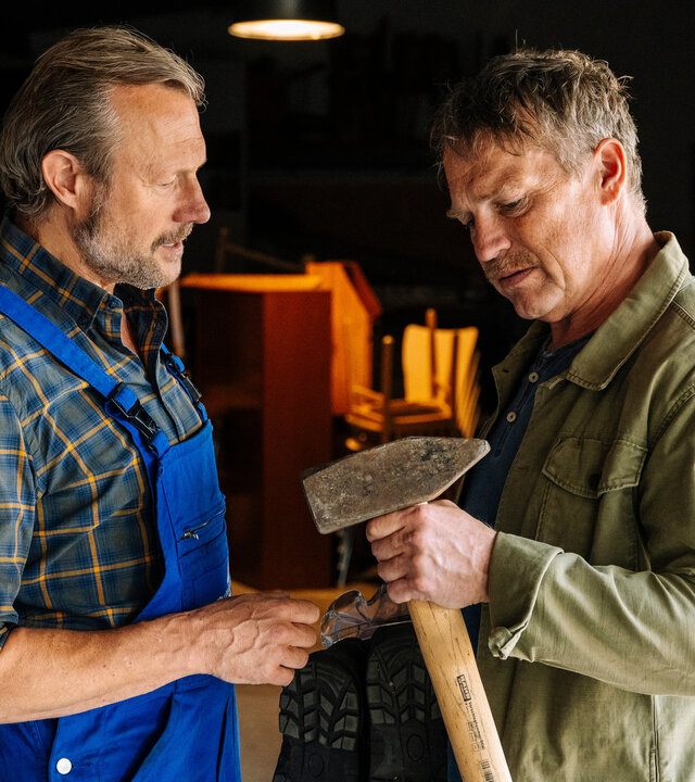 "SOKO Köln - Unvergessen": Matti Wagner (Pierre Besson) steht in einem Lagerraum und hält eine Axt in der Hand, die er nachdenklich betrachtet. Carlo Kühn (Hanno Friedrich) steht nah vor ihm und blickt ebenfalls auf die Axt. Er trägt eine blaue Latzhose über einem karierten Hemd. Im Hintergrund stehen Regale, Kartons und Werkstattausrüstung.
