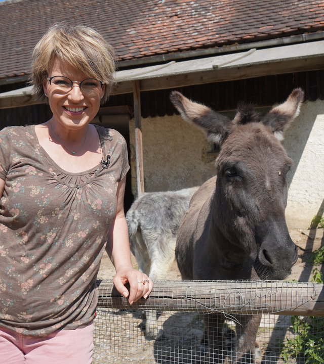 "sonntags - Öko = anders = gut?": Andrea Ballschuh steht vor einem Esel im Gehege. Sie lehnt sich an den Holzzaun und lächelt in die Kamera. Der Esel steht neben ihr hinter dem Gatter und schaut ebenfalls in die Kamera.