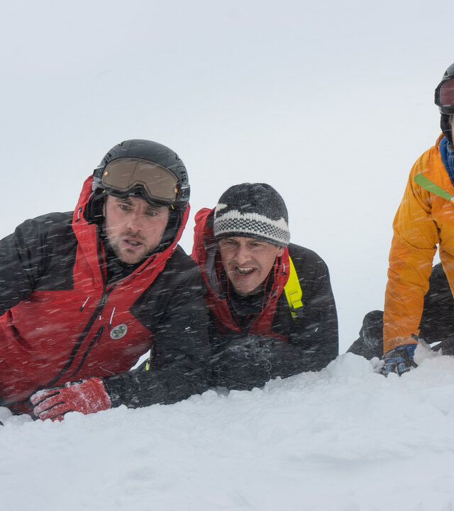 "Die Bergretter: Eiskalte Wahrheit": Simon (Ferdinand Seebacher), Rudi (Michael Pascher) und Markus (Sebastian Ströbel) liegen in voller Montur im Schnee, ihre Blicke sind bestürzt. Es schneit.