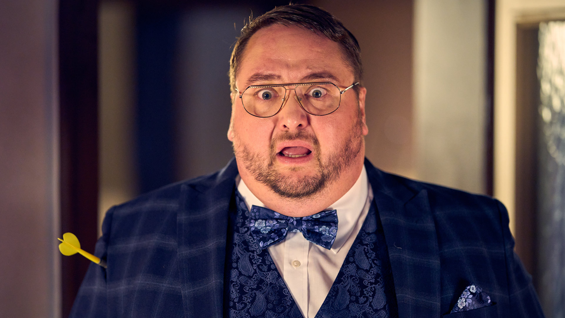 "Ich dich auch! - Der Pfeil": Tüffi (Daniel Zillmann) steht schick angezogen wie unter Schock mit aufgerissenen Augen und Mund im Zimmer. Ein Dartpfeil hat ihn am Arm getroffen.