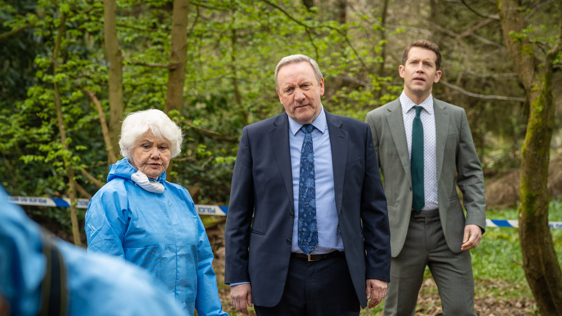 "Inspector Barnaby - Das Buch Bertram": Fleur Perkins (Annette Badland), Detective Chief Inspector John Barnaby (Neil Dudgeon und Detective Constable Jamie Winter (Nick Hendrix) stehen nebeneinander im Wald. Hinter ihnen ist ein Absperrband der Polizei gespannt.