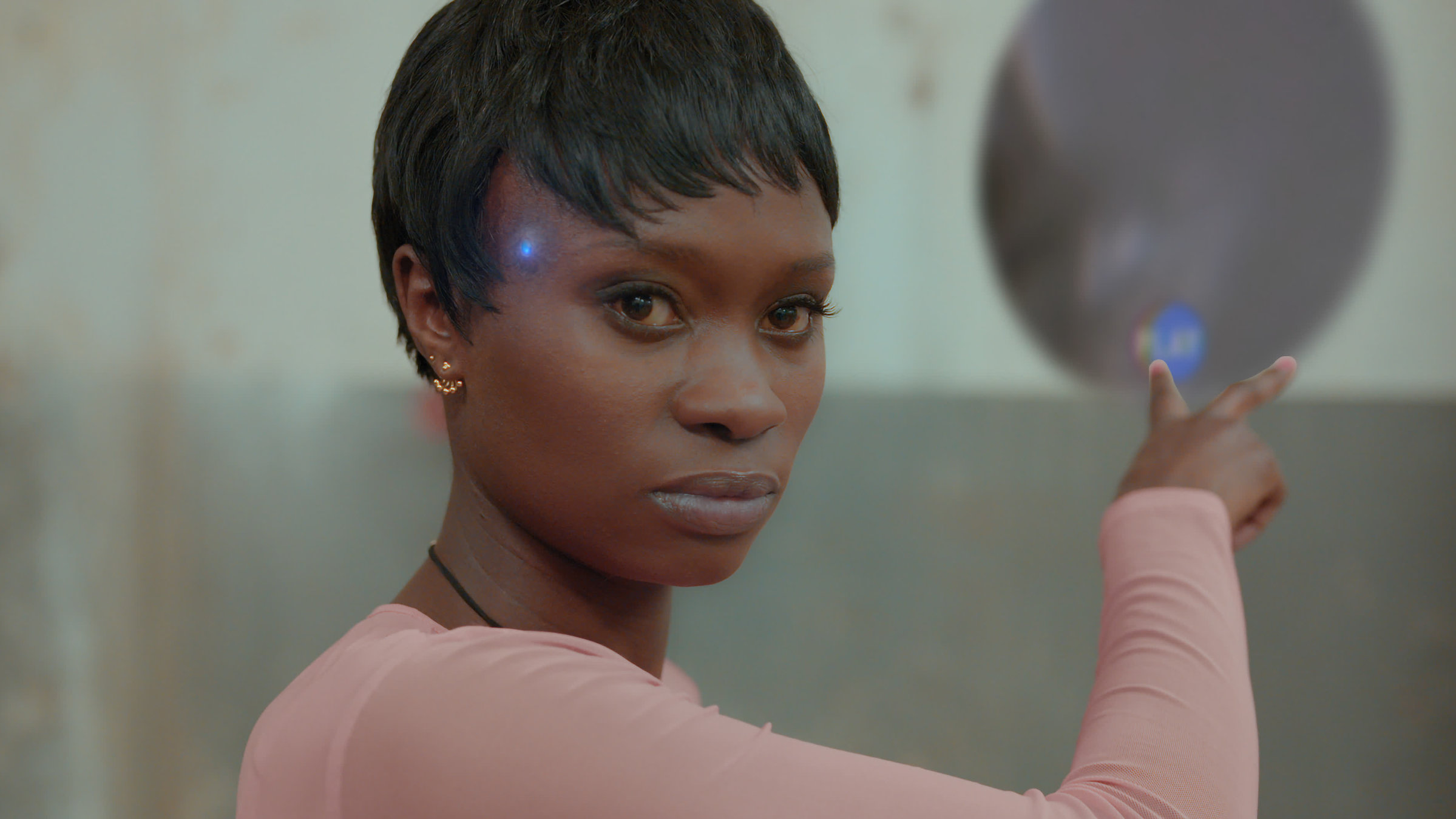 "Hyperland": Cee (Lorna Ishema) zeigt mit ihrem Zeige- und Mittelfinger auf eine hinter ihr liegende Wand. An ihrer Schläfe leuchtet eine blaue Leuchtdiode.