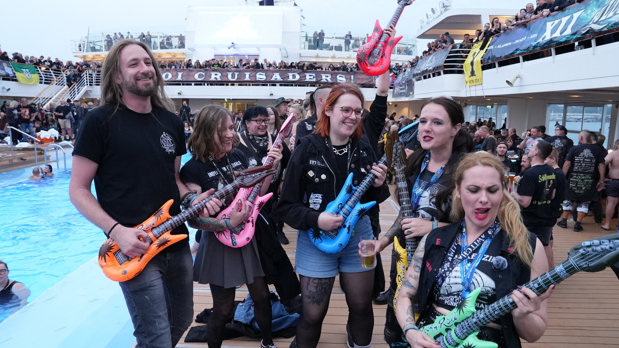"ZDF.reportage: Heavy Metal Kreuzfahrt": Im Vordergrund eine Gruppe von fünf jungen Heavy Matal Fans, die mit aufblasbaren Spielzeuggitarren Spaß haben. Im Hintergrund liegt der Pool und das Oberdeck des Kreuzfahrtschiffes. Alle Gänge sind voller Menschen.