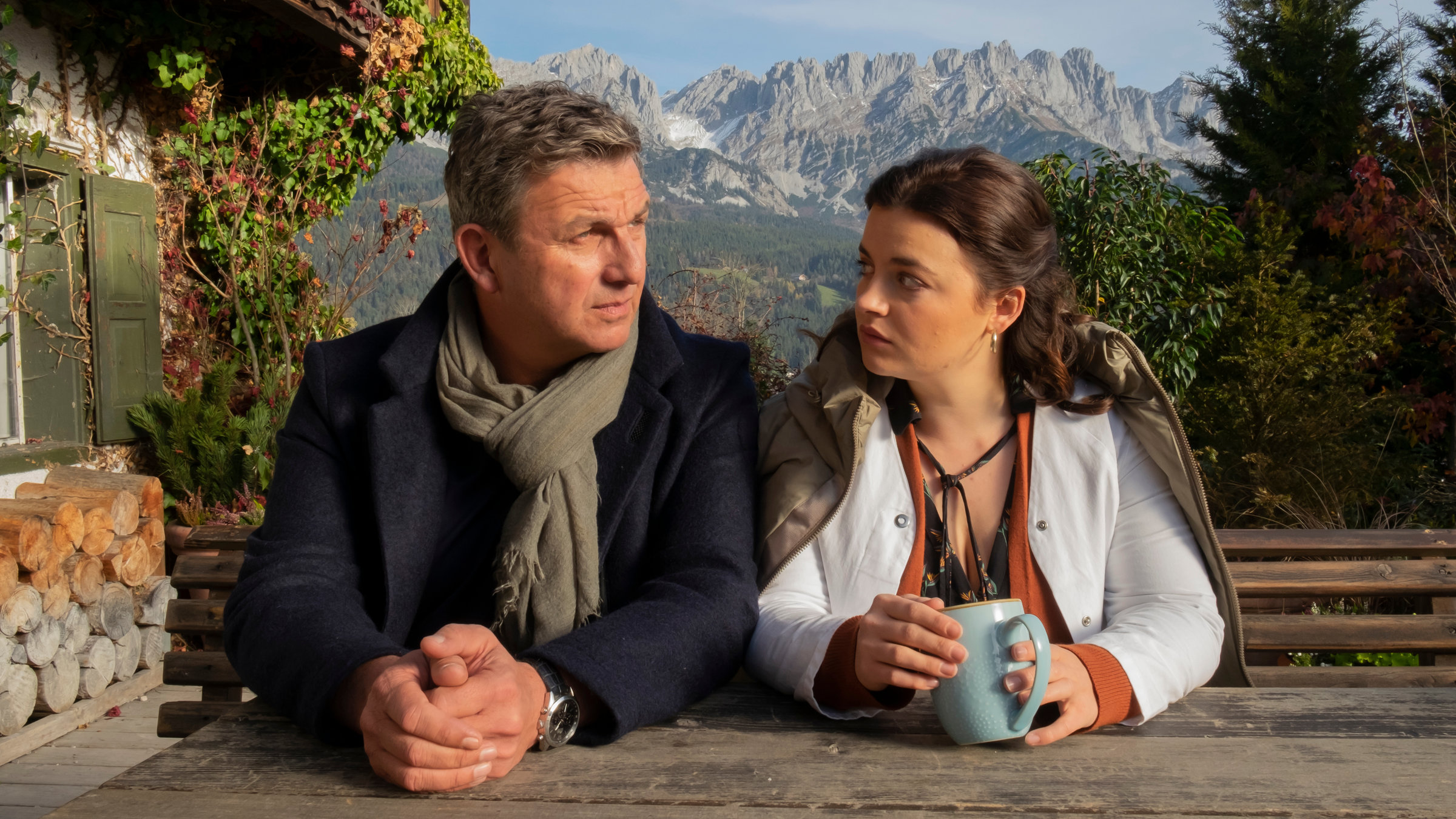 "Der Bergdoktor: Was bleibt": Martin (Hans Gruber) und Lilli (Ronja Forcher) sitzen nebeneinander vor der Praxis auf der Bank und blicken sich nachdenklich an. Im Hintergrund eine Bergkulisse.