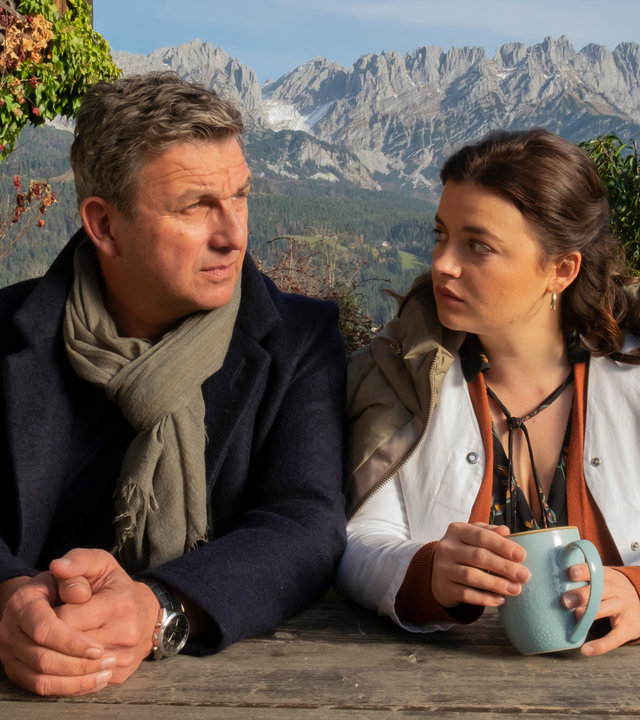 "Der Bergdoktor: Was bleibt": Martin (Hans Gruber) und Lilli (Ronja Forcher) sitzen nebeneinander vor der Praxis auf der Bank und blicken sich nachdenklich an. Im Hintergrund eine Bergkulisse.
