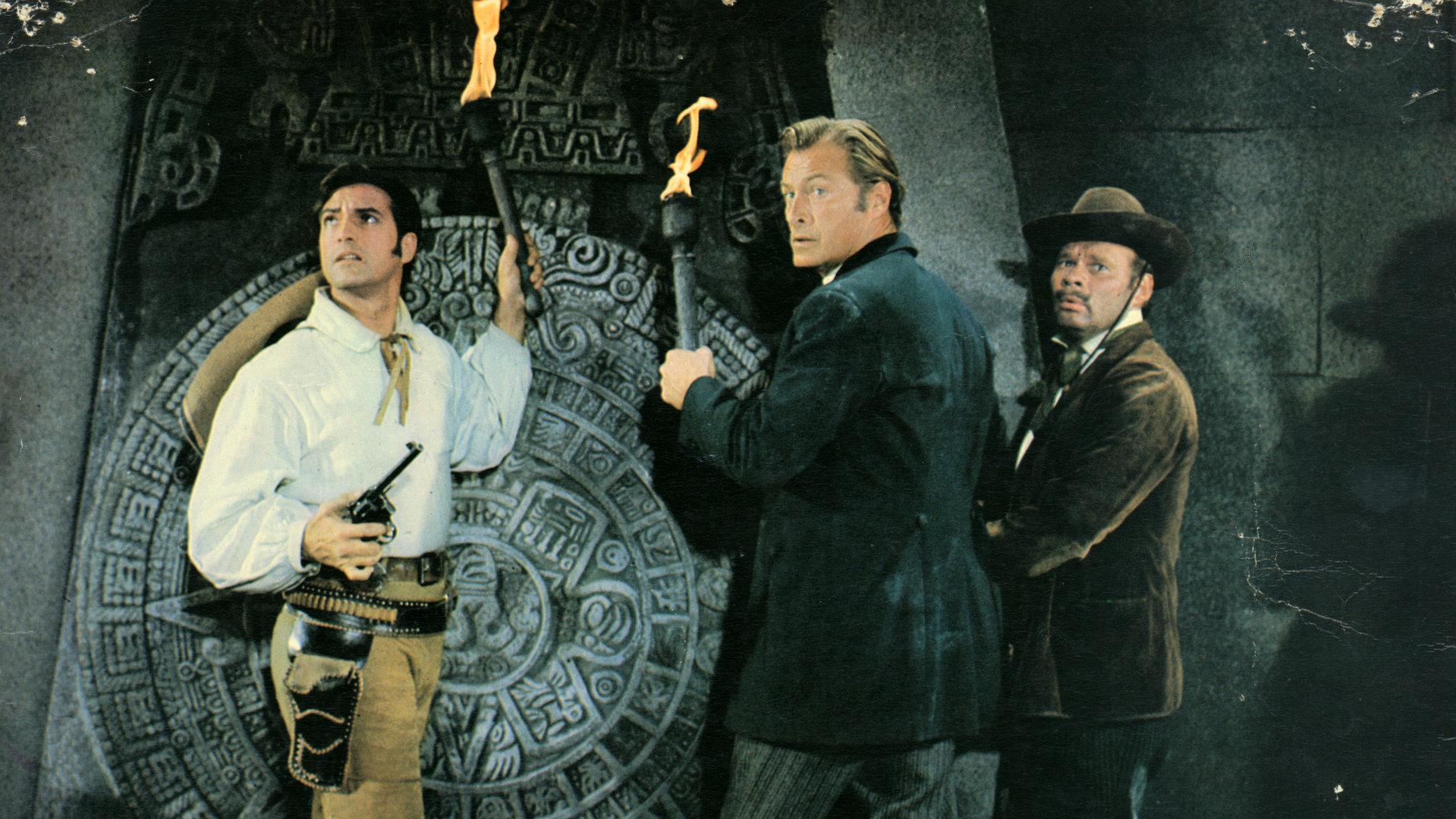 "Die Pyramide des Sonnengottes": Dr. Karl Sternau (Lex Barker), Graf Alfonso (Gérard Barray) und André Hasenpfeffer (Ralf Wolter) haben Fackeln in den Händen und schauen sich um. Sie stehen vor einem riesigen steinernen Inka-Ornament.