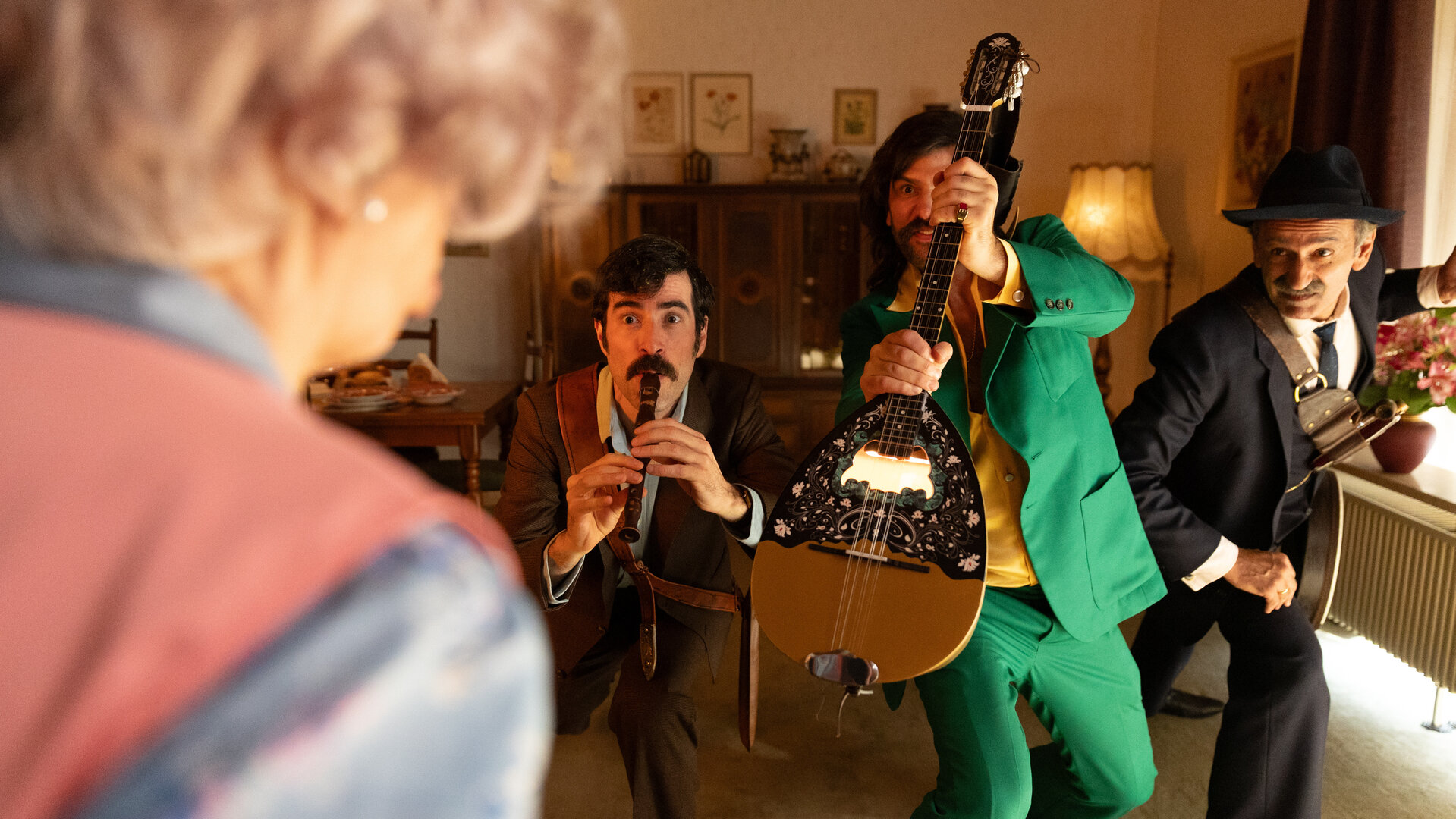 "Rembetis - die Geisterjäger - Maneater": Hektor (Samy Challah) steht gemeinsam mit Paris (Jasin Challah) und Marcos (Pavlos Kourtidis) in einem Wohnzimmer. Hektor spielt auf einer Flöte, Paris hält eine Bouzouki, während Marcos sich nach vorne lehnt. Vor ihnen steht, verschwommen von hinten zu sehen, Marie Meierling (Johanna Gastdorf).