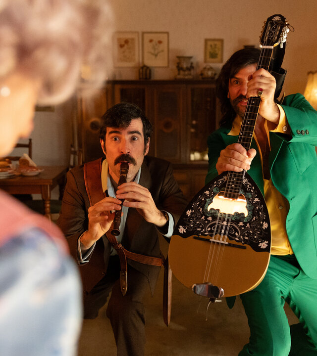 "Rembetis - die Geisterjäger - Maneater": Hektor (Samy Challah) steht gemeinsam mit Paris (Jasin Challah) und Marcos (Pavlos Kourtidis) in einem Wohnzimmer. Hektor spielt auf einer Flöte, Paris hält eine Bouzouki, während Marcos sich nach vorne lehnt. Vor ihnen steht, verschwommen von hinten zu sehen, Marie Meierling (Johanna Gastdorf).