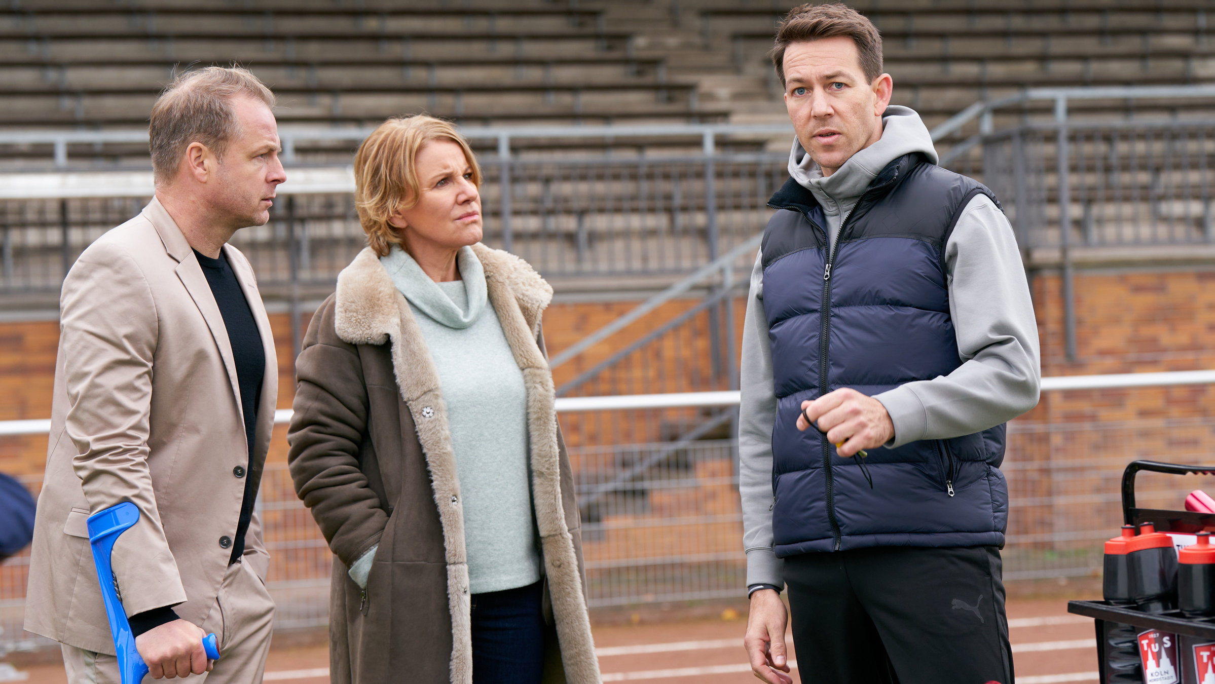 "Marie Brand und die falsche Wahrheit": Marie Brand (Mariele Millowitsch) und Jürgen Simmel (Hinnerk Schönemann) stehen auf dem Sportplatz und schauen den Trainer Victor Holtmann (Florian Anderer) an. Victor Holtmann hält in seiner linken Hand eine Pfeife und schaut links aus dem Bild heraus. Jürgen Simmel stützt sich mit seiner rechten Hand an einer Krücke ab.