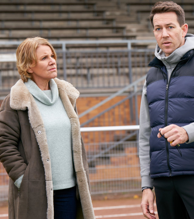 "Marie Brand und die falsche Wahrheit": Marie Brand (Mariele Millowitsch) und Jürgen Simmel (Hinnerk Schönemann) stehen auf dem Sportplatz und schauen den Trainer Victor Holtmann (Florian Anderer) an. Victor Holtmann hält in seiner linken Hand eine Pfeife und schaut links aus dem Bild heraus. Jürgen Simmel stützt sich mit seiner rechten Hand an einer Krücke ab.