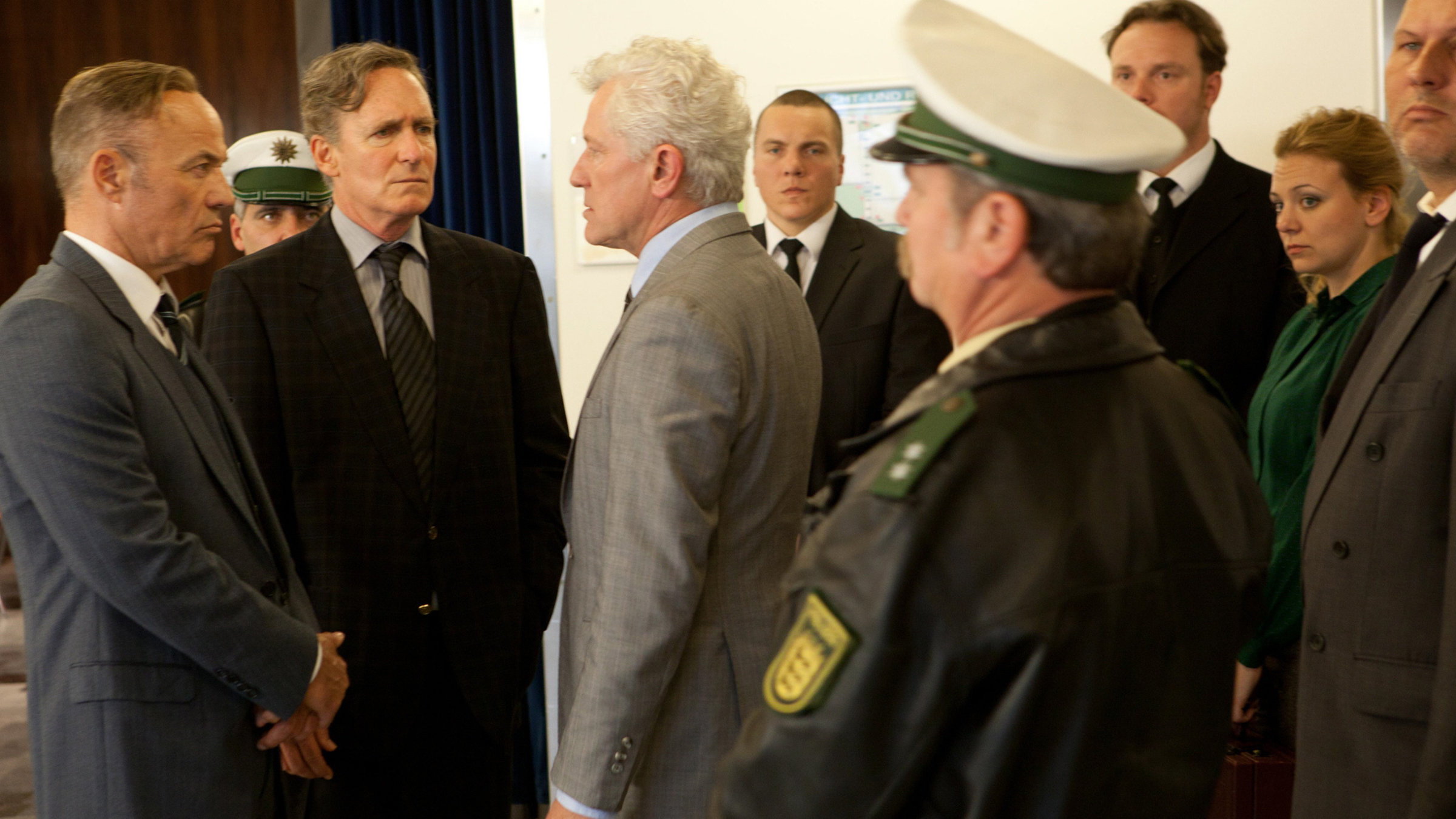 "Der blinde Fleck - Das Oktoberfestattentat": Von links: der Leiter des bayerischen Staatsschutzes, Dr. Hans Langemann (Heiner Lauterbach), dessen rechte Hand Meier (August Zirner) und Generalbundesanwalt Kurt Rebmann (Miroslav Nemec) stehen sich gegenüber und schauen sich an. Hinter ihnen stehen zwei Polizeibeamte und weitere Menschen.