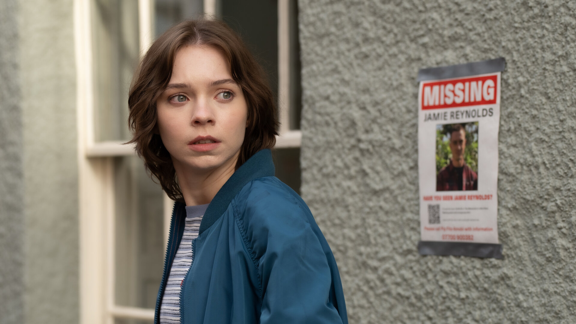 "A Good Girl’s Guide to Murder – Das Verschwinden von Jamie Reynolds": Pip Fitz-Amobi (Emma Myers) steht in einer Gasse neben einem "Missing"-Poster von Jamie Reynolds. Sie schaut angespannt zur Seite.