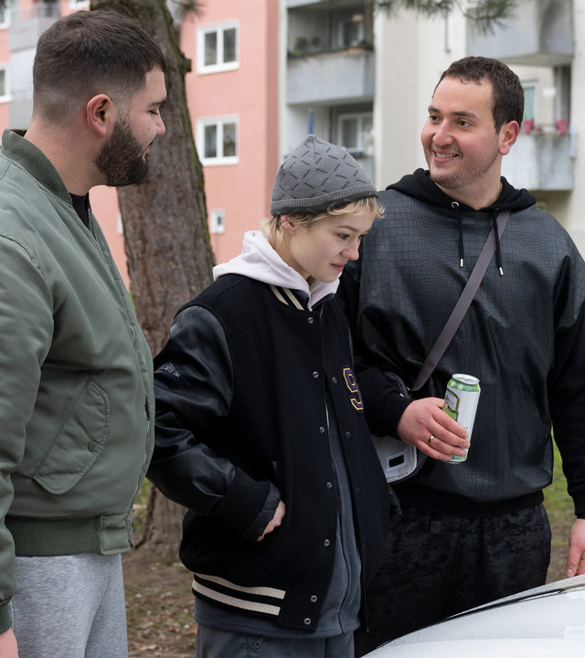 "Becoming Charlie - Gegen den Strom": Nikolas (Danilo Kamperidis), Charlie (Lea Drinda) und Enzo (Rouven Israel) stehen nebeneinander vor dem Kühler eines weißen Fahrzeugs und unterhalten sich. Während Charlie lächelnd auf den Kühler blickt, sehen sich Nikolas und Enzo lachend an. Enzo hält eine Getränkedose in der rechten Hand. Im Hintergrund ist eine Wohnanlage mit Hochhäusern zu sehen.