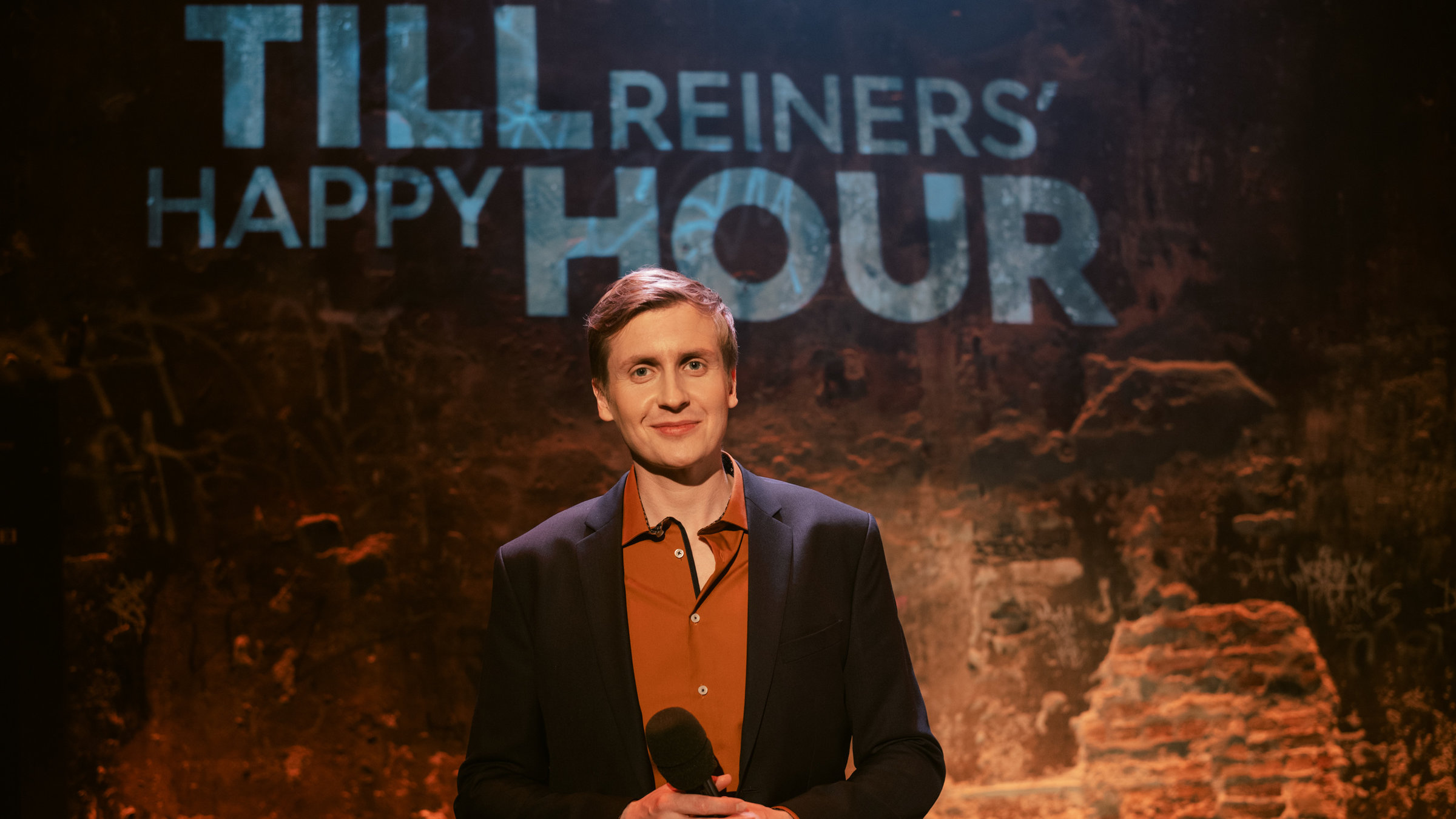 "Till Reiners‘ Happy Hour": Till Reiners steht mit einem Mikrofon in der Hand auf der Bühne.