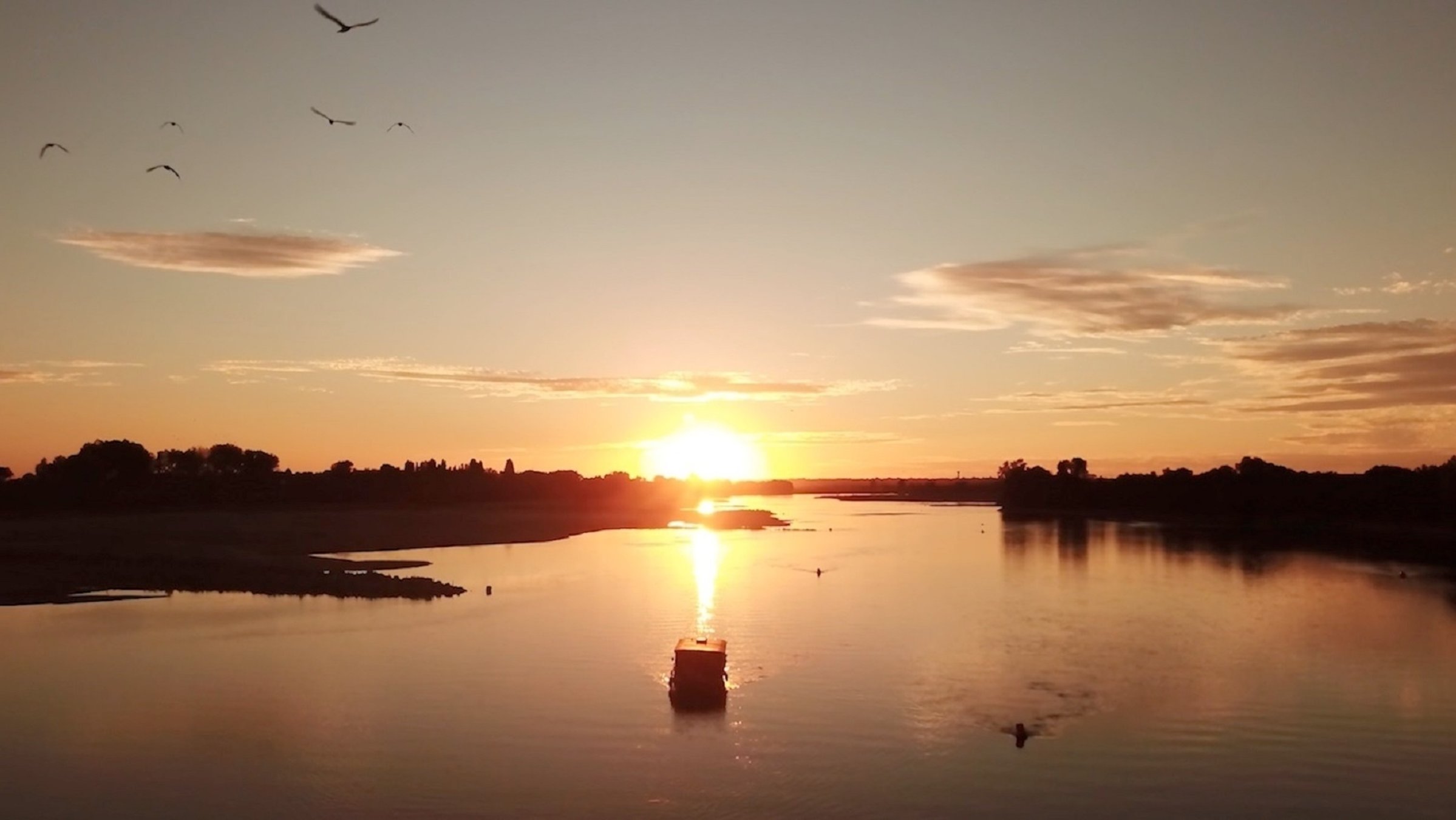 "Das Loiretal - Von Schloss zu Schloss": Panoramaaufnahme der Loire mit ihrem breiten Flussbett: Die untergehende Sonne taucht die Landschaft in rötliches Licht. Am Himmel hängen einige Wolken. In der Mitte des Flusses fährt ein Boot.
