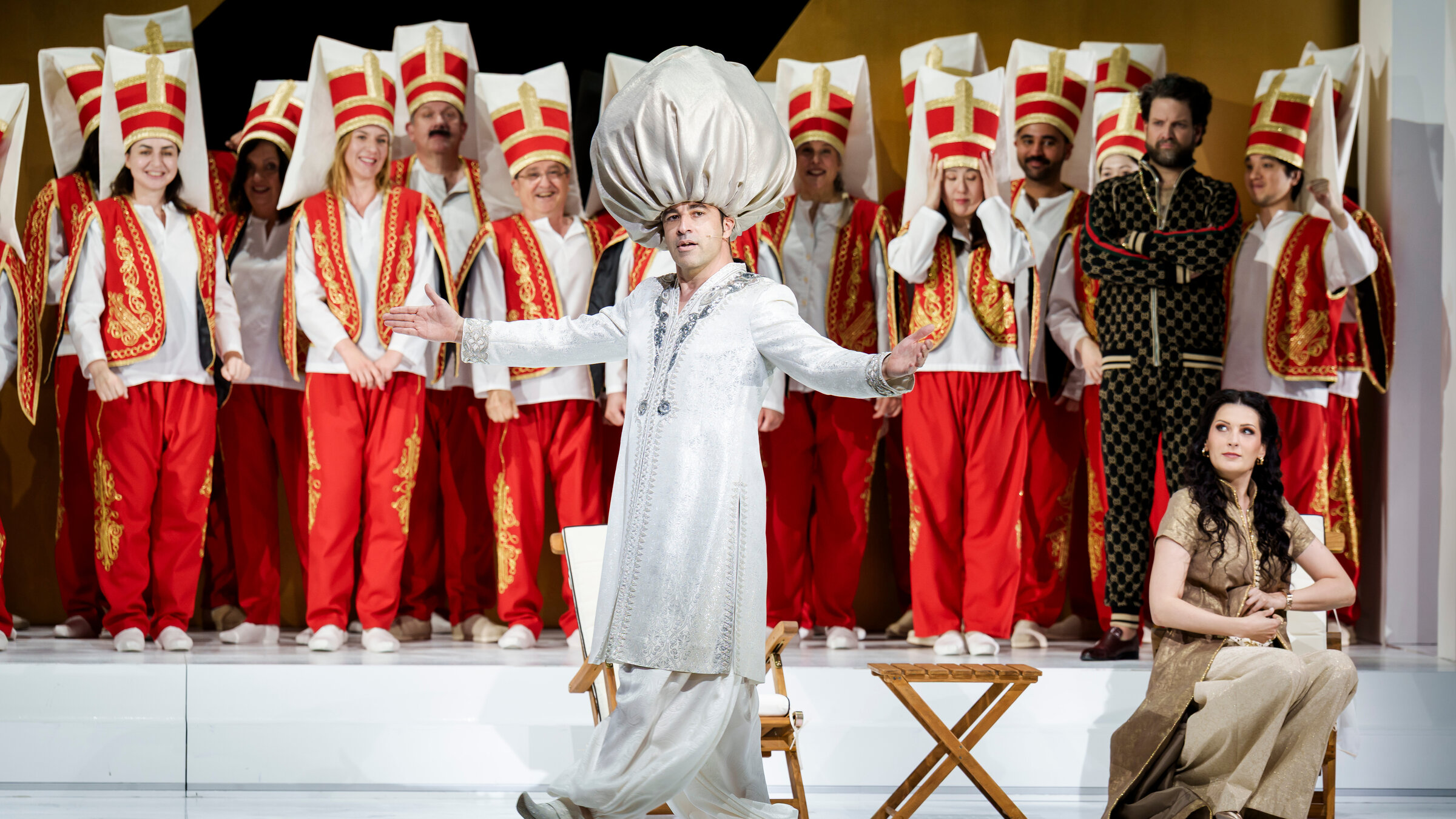"Die Entführung aus dem Serail": Mittig auf einer Bühne steht Bassa Selim (gespielt von Bülent Ceylan). Er ist bekleidet mit einem weißen Kaftan und einem übergroßen weißen Turban. Er hat die Arme ausgestreckt. Rechts im Bild sitzt eine Frau auf einem Stuhl und blickt in Richtung von Bassa Selim. Dahinter stehen etwas erhöht zahlreiche Menschen. Sie alle sind einheitlich gekleidet, mit roten besticken Hosen, einem weißen Oberteil und einer roten bestickten Weste. Alle tragen hohe Hüte in Rot, Gold und weiß.