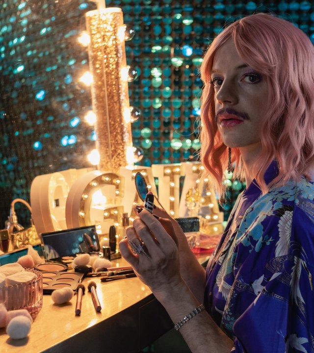"SOKO Hamburg - Glamour, Girls!": Paul Siebert alias Gaia Glamour (Merlin Sandmeyer) sitzt vor einem beleuchteten Spiegel, der auf einer Schminkkommode steht und blickt ernst nach links. Er trägt einen blau-gemusterten Kimono und eine rosafarbene Perücke. Vor ihm auf der Kommode liegen diverse Kosmetikartikel. Im Hintergrund sieht man einen blauen Pailettenvorhang.