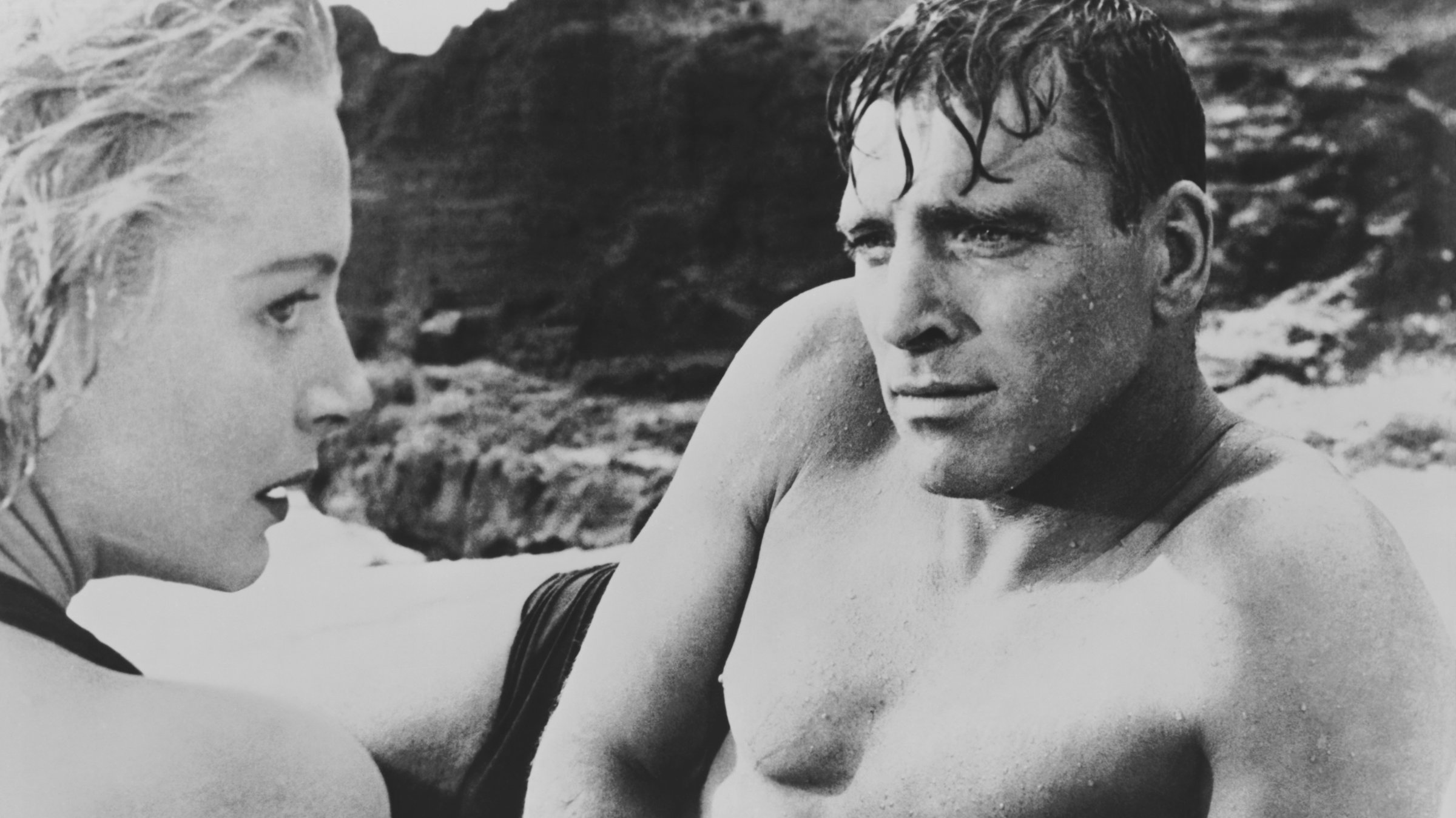 "Verdammt in alle Ewigkeit": Karen Holmes (Deborah Kerr) und Sergant Milton Warden (Burt Lancaster) sitzen einander in Badekleidung und mit nassen Haaren an einem Strand gegenüber. Im Hintergrund sind Felsen zu sehen. Er schaut sie ernst an, sie weicht seinem Blick aus.