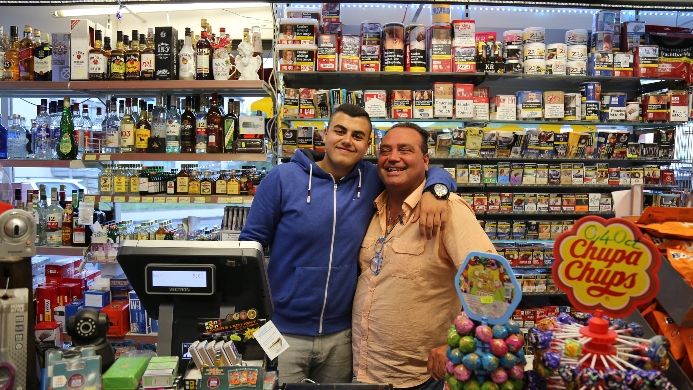 "ZDF.reportage: Treffpunkt Kiosk - Wo das Herz des Viertels schlägt": Yusuf steht mit seinem Vater und Inhaber Vahit Arm in Arm hinter der Kiosk-Kasse.