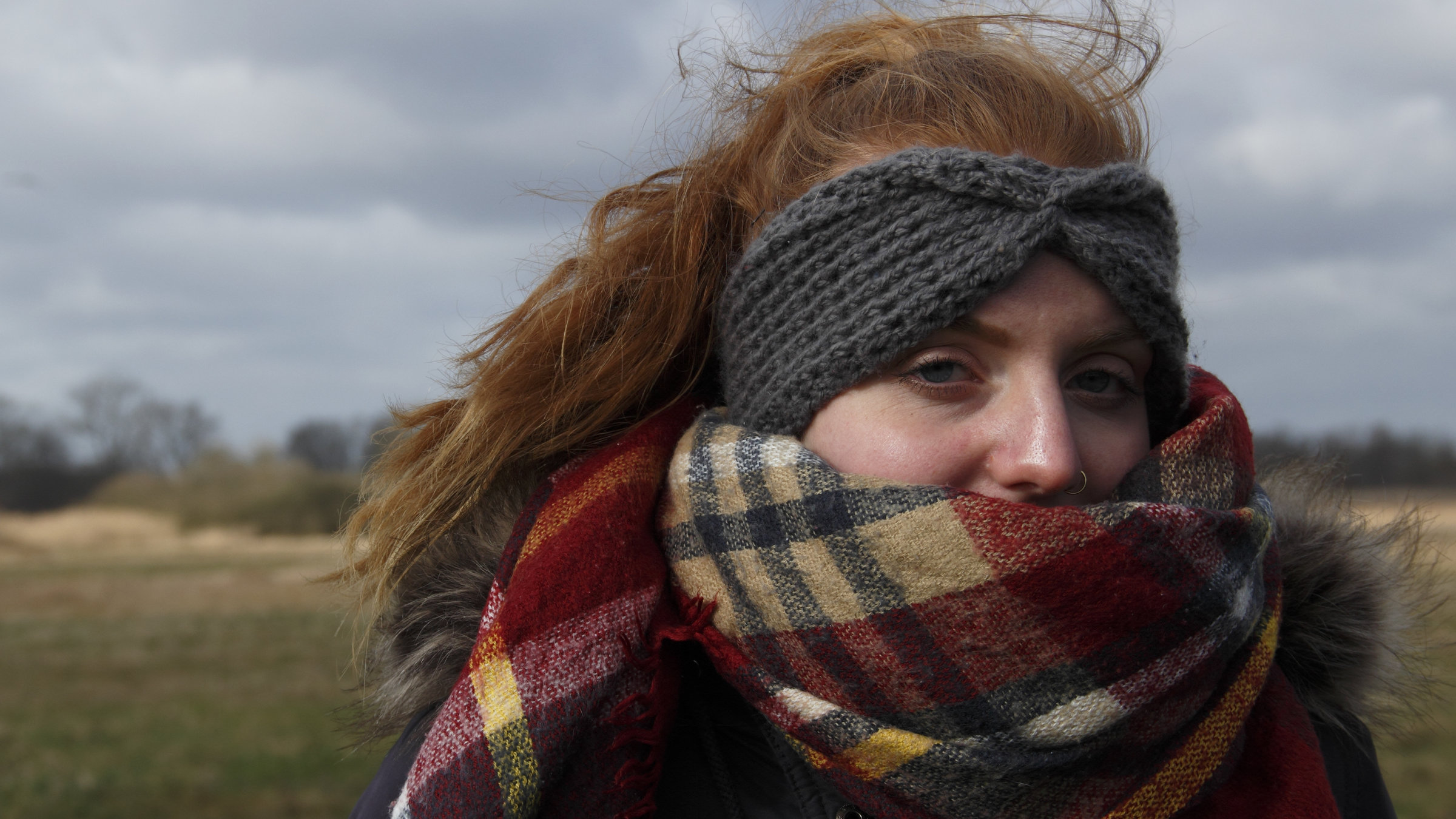 "37°: Ich bin noch da - Suizidgedanken junger Menschen": Junge rothaarige Frau mit langem Zopf und einem grauen Wollstirnband, das Gesicht ein Stück weit von einem Schal bedeckt, steht in einer Naturlandschaft.