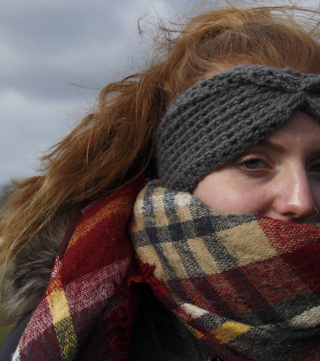 "37°: Ich bin noch da - Suizidgedanken junger Menschen": Junge rothaarige Frau mit langem Zopf und einem grauen Wollstirnband, das Gesicht ein Stück weit von einem Schal bedeckt, steht in einer Naturlandschaft.