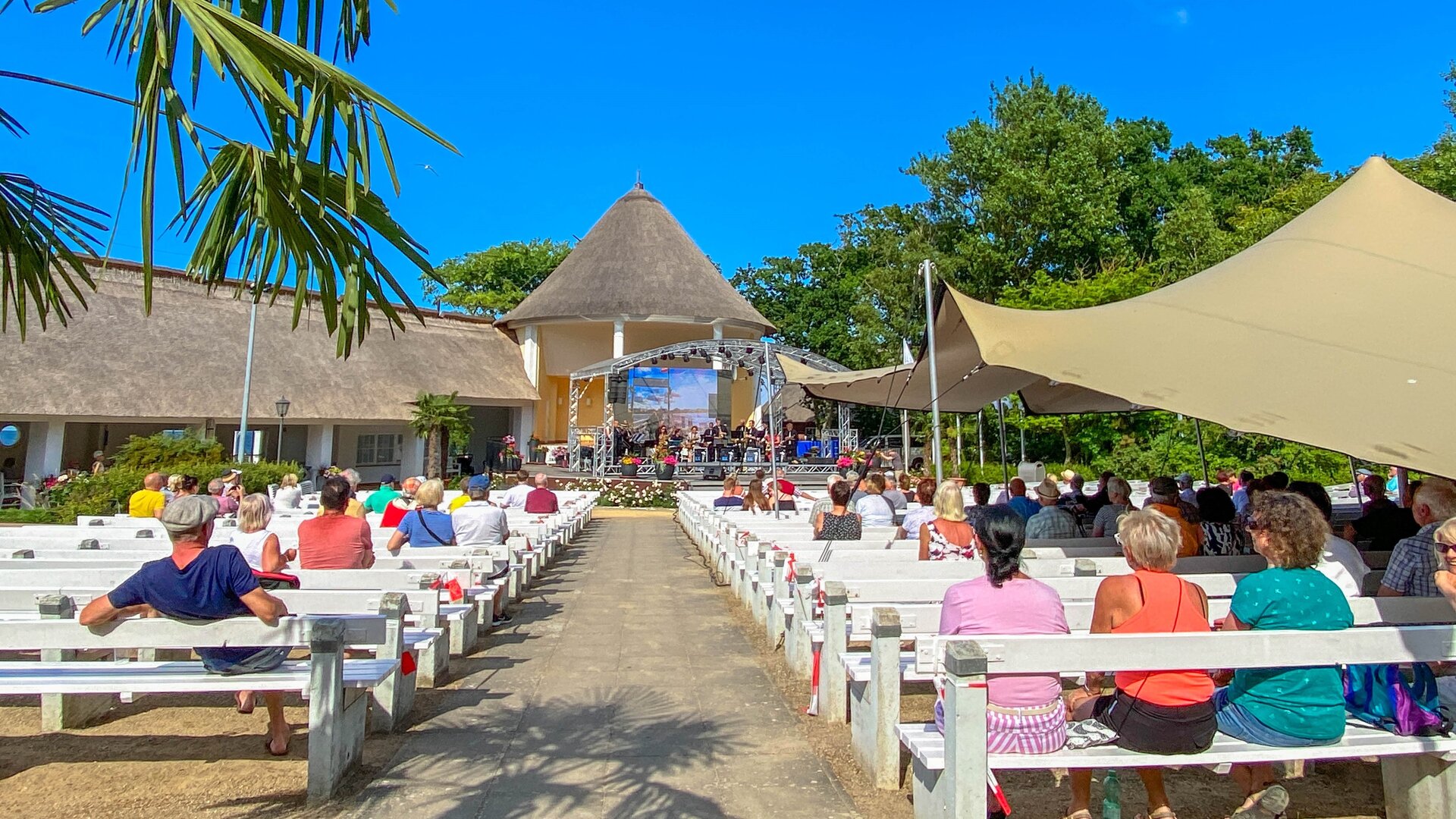 "Evangelischer Gottesdienst - Lass gut sein": Im Konzertgarten des Ostseebads Kühlungsborn führt ein Weg zwischen weißen Holzbänken hindurch direkt zu einer Bühne. Auf den Bänken sitzen Zuhörende unter Sonnensegeln, der Himmel leuchtet tiefblau.