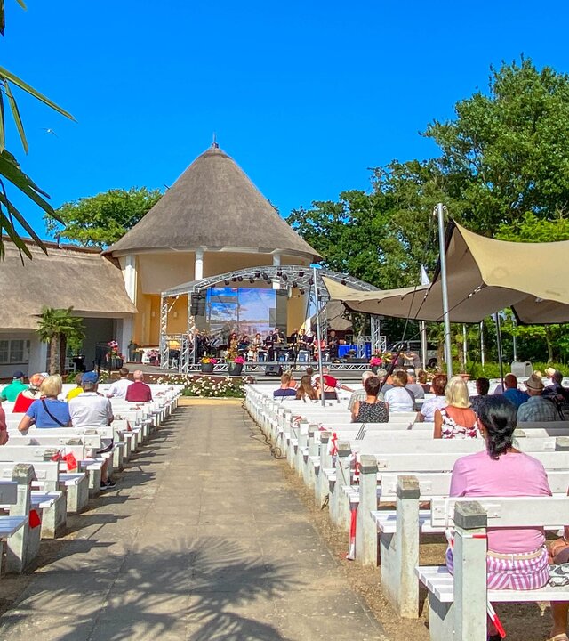 "Evangelischer Gottesdienst - Lass gut sein": Im Konzertgarten des Ostseebads Kühlungsborn führt ein Weg zwischen weißen Holzbänken hindurch direkt zu einer Bühne. Auf den Bänken sitzen Zuhörende unter Sonnensegeln, der Himmel leuchtet tiefblau.