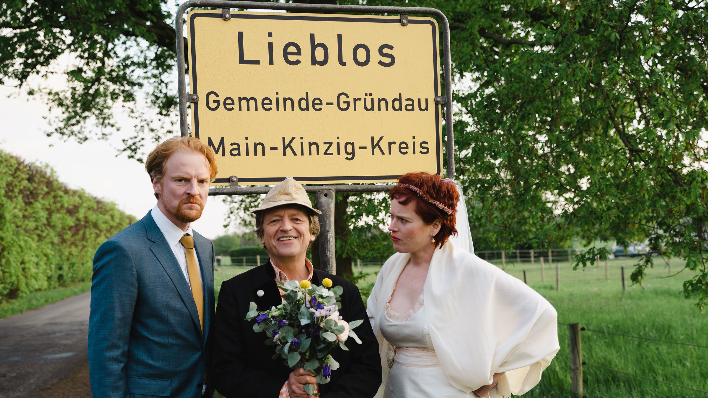 "Beim Pelzig auf der Bank - Scheiß Liebe": Pelzig steht vor dem Ortsschild von "Lieblos" und hält einen Blumenstrauß in den Händen. Auf seiner linken Seite ein Bräutigam, auf seiner rechten Seite eine Braut. Beide starren ihn verärgert an.