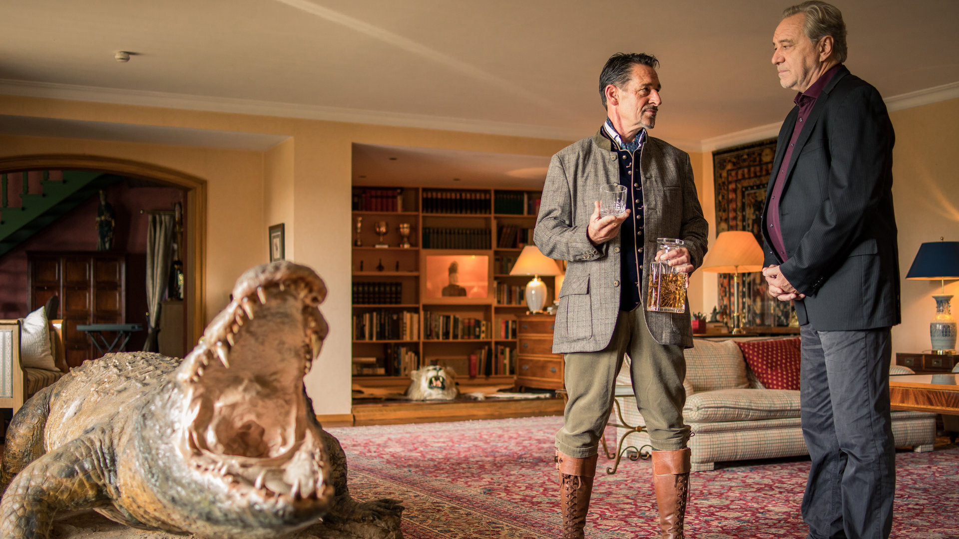 "SOKO München - Isegrim": Arthur Bauer (Gerd Silberbauer) steht mit Elmar Dannenberg (Max Tidof) in dessen Wohnzimmer. Arthur Bauer sieht Elmar Dannenberg ernst an und dieser blickt zurück. Elmar Dannenberg trägt seine Jagdklamotten und hält in der rechten Hand ein leeres Glas. In seiner linken Hand befindet sich eine Cognac Flasche. Im hinteren Teil des großen Wohnraumes sieht man ein Bücherregal und einen Tigerfell Teppich . Außerdem befindet sich ein beiges Sofa und drei Kommoden im Raum. Auf jeder Kommode steht eine Stehlampen, die warmes Licht spendet. Vor den beiden Männern steht eine lebensgroße Krokodilfigur mit aufgerissenem Maul.