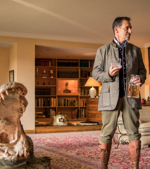 "SOKO München - Isegrim": Arthur Bauer (Gerd Silberbauer) steht mit Elmar Dannenberg (Max Tidof) in dessen Wohnzimmer. Arthur Bauer sieht Elmar Dannenberg ernst an und dieser blickt zurück. Elmar Dannenberg trägt seine Jagdklamotten und hält in der rechten Hand ein leeres Glas. In seiner linken Hand befindet sich eine Cognac Flasche. Im hinteren Teil des großen Wohnraumes sieht man ein Bücherregal und einen Tigerfell Teppich . Außerdem befindet sich ein beiges Sofa und drei Kommoden im Raum. Auf jeder Kommode steht eine Stehlampen, die warmes Licht spendet. Vor den beiden Männern steht eine lebensgroße Krokodilfigur mit aufgerissenem Maul.