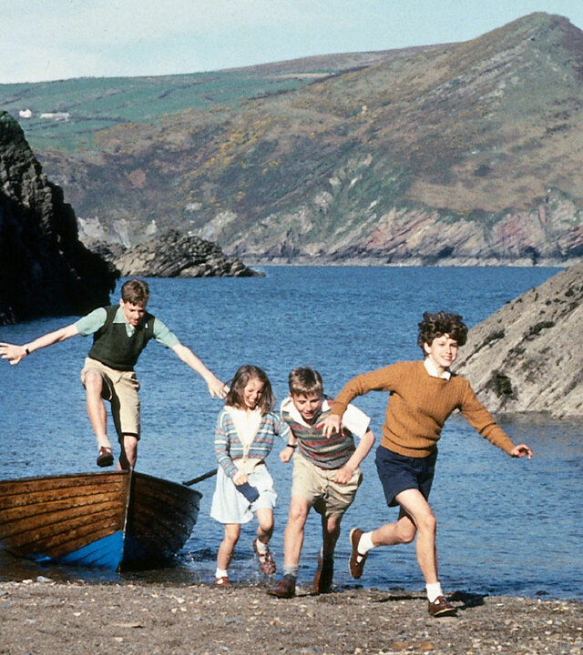 "Fünf Freunde erforschen die Schatzinsel": Julian (Marco Williamson), Anne (Laura Petela), Dick (Paul Child) und George (Jemima Rooper) springen lachend an einem felsigen Ufer aus einem Ruderboot und rennen in Richtung Strand, während der Hund Timmy neben ihnen herläuft.
