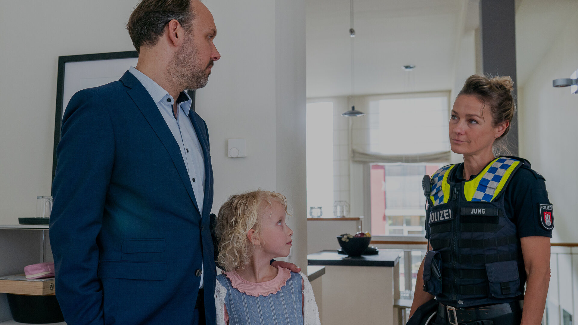 "Notruf Hafenkante - Tierwohl": Bernd Schröter (Felix Knopp) steht neben seiner Tochter Lara (Käthe-Marie Meyer) in der Küche. Er hat seine linke Hand auf Laras rechten Schulter gelegt. Beide blicken zu der Polizistin Franzi Jung (Rhea Harder-Vennewald), die seitlich neben ihnen steht. Franzi schaut Bernd Schröter nachdenklich an.