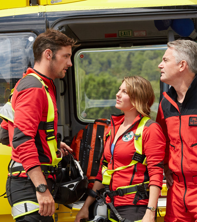 "Die Bergretter: Tod am Dachstein": Der Rettungshubschrauber steht am Hangar mit geöffneten Türen. Davor stehen, in voller Montur, Simon (Ferdinand Seebacher), Katharina (Luise Bähr) und Michael (Robert Lohr) und unterhalten sich.