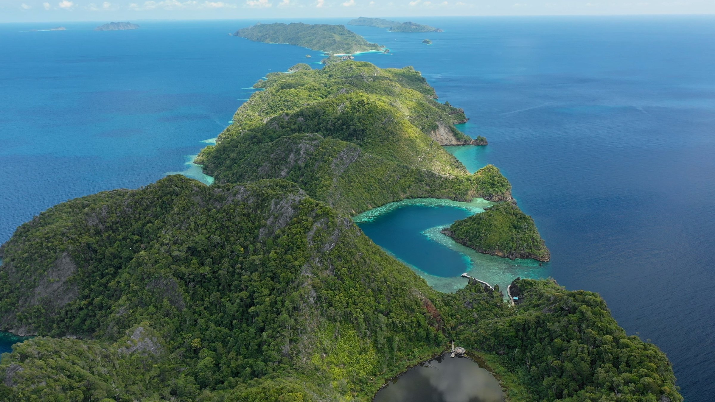 "Into the Blue – Indonesiens Unterwasserparadiese - Im Archipel Raja Ampat": Luftbild einer grünen Insel mit herzförmiger Lagune. Im Hintergrund Meer mit einzelnen Inseln.