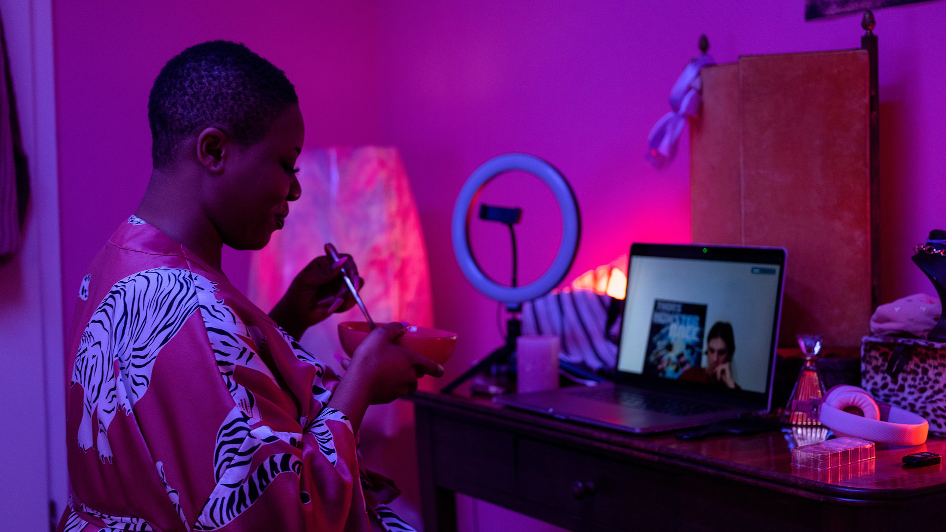 "WatchMe - Sex sells - Invest in yourself!": Malaika (Maddy Forst) sitzt in einem lila beleuchteten Zimmer an einem Schminktisch, auf dem ein Laptop steht. Sie hält eine Schüssel und einen Löffel in ihren Händen. Sie trägt einen Morgenmantel aus Satin. Ihr gegenüber, auf dem Bildschirm des Laptops, ist Kai3000 zu sehen.