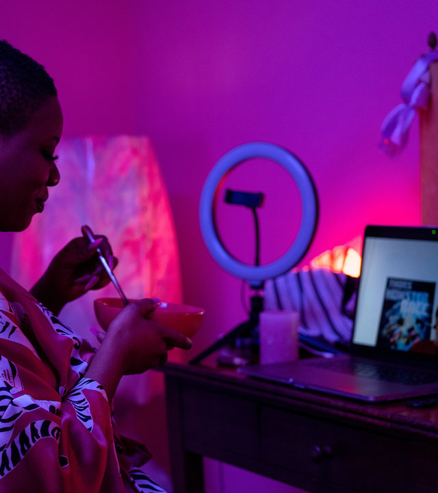 "WatchMe - Sex sells - Invest in yourself!": Malaika (Maddy Forst) sitzt in einem lila beleuchteten Zimmer an einem Schminktisch, auf dem ein Laptop steht. Sie hält eine Schüssel und einen Löffel in ihren Händen. Sie trägt einen Morgenmantel aus Satin. Ihr gegenüber, auf dem Bildschirm des Laptops, ist Kai3000 zu sehen.