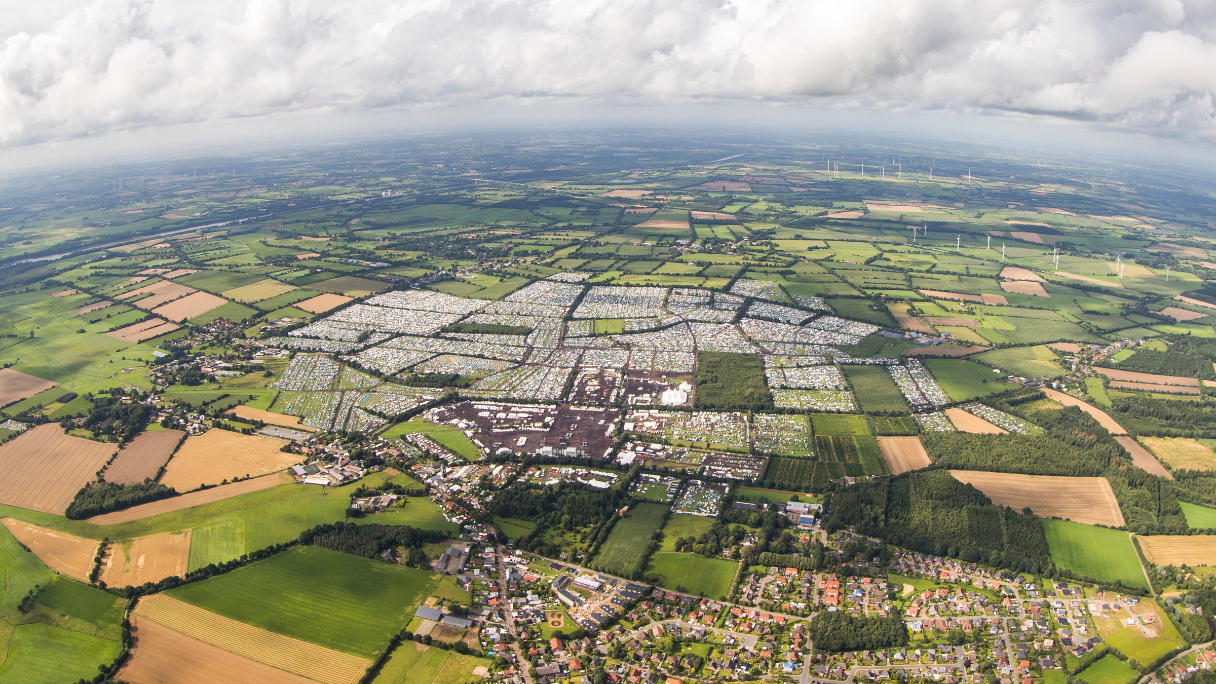 "Wacken 2019": Luftaufnahme des gesamten Geländes vom „Wacken Open Air“. Zu sehen sind die Campingplätze, die Bühnen und das gesamte Festivalareal, das direkt neben dem Dorf Wacken liegt. Außen herum sind viele Wiesen und Natur.